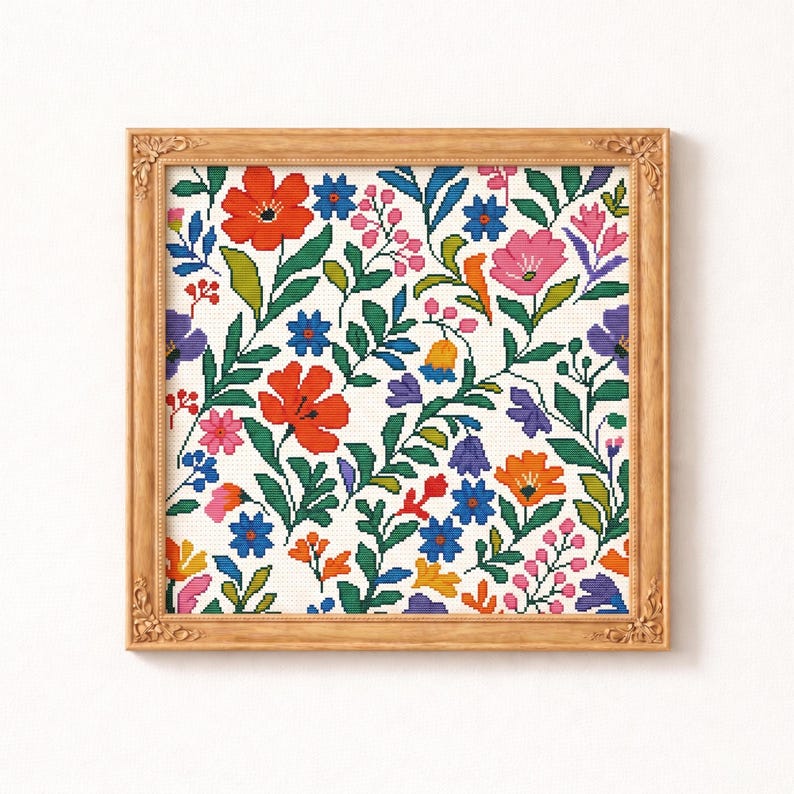 May include: A framed floral cross-stitch pattern. The design features a variety of colorful flowers, including red, pink, orange, and blue, with green leaves against a white background. The frame is a light brown color with ornate detailing.