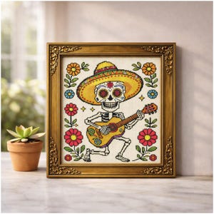 May include: Cross-stitch art of a skeleton playing a guitar, wearing a sombrero. The skeleton is surrounded by colourful flowers and framed in a gold ornate frame. The artwork is displayed on a white surface.