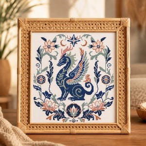 May include: A square cross-stitch embroidery featuring a central blue dragon with wings, surrounded by a symmetrical floral border in shades of blue, peach, and sage green, encased in a light-coloured wooden frame with a woven cane border.