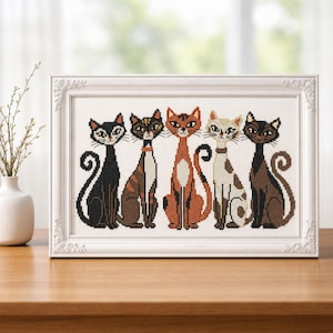 Puede incluir: Un bordado a punto de cruz enmarcado con cinco gatos estilizados. Los gatos son de varios colores, incluyendo negro, marrón y naranja, con collares decorativos. El marco es blanco con detalles ornamentados, sobre una superficie de madera.