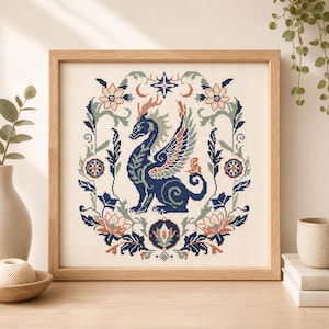 May include: A square cross-stitch embroidery in a light wood frame featuring a dark blue dragon with orange and light blue wings, centred within a symmetrical floral wreath of sage green, peach, and dark blue botanical elements on cream fabric.