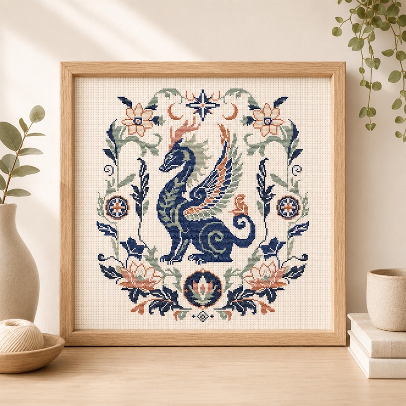 May include: A square cross-stitch embroidery in a light wood frame featuring a dark blue dragon with orange and light blue wings, centred within a symmetrical floral wreath of sage green, peach, and dark blue botanical elements on cream fabric.