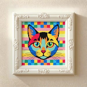 Puede incluir: Una obra de arte en punto de cruz enmarcada que representa la cara de un gato colorido. El gato está representado en azul, amarillo, naranja, rosa y negro. El fondo es una mezcla de rayas horizontales y cuadrados coloridos.