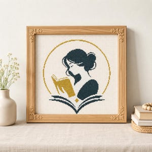 May include: A square cross-stitch embroidery in a light wood frame featuring a dark blue silhouette of a person reading a gold book. The design is enclosed within a thin gold circular border, set against a white fabric background with a grid pattern.