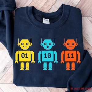 Könnte beinhalten: Marineblaues Sweatshirt mit drei bunten Roboter-Designs. Die Roboter sind gelb, blau und orange, jeder mit einer anderen Nummer: 01, 10 und 11. Das Design ist auf einem bequemen Sweatshirt gedruckt.