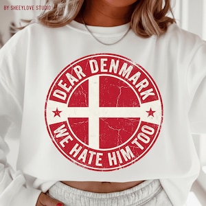 Könnte beinhalten: Weißes Sweatshirt mit einem verblassten rot-weißen Grafikdesign. Das Design ist ein kreisförmiges Design mit den Worten "DEAR DENMARK WE HATE HIM TOO" um ein weißes Kreuz auf rotem Hintergrund. Zwei rote Sterne auf beiden Seiten des Kreuzes.