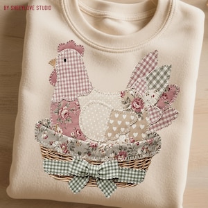 May include: Cream-colored sweatshirt featuring a patchwork chicken design. The chicken is composed of various patterned fabrics, including gingham, floral, and polka dots, and sits in a woven basket with a green gingham bow. The text "BY SHEEYLOVE STUDIO" is in the upper left corner.