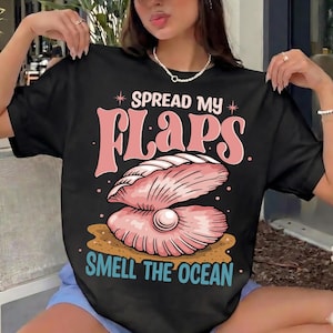 以下が含まれることがあります： ピンク色の貝殻のグラフィックと「SPREAD MY FLAPS SMELL THE OCEAN」のテキストが入った黒いTシャツ。デザインには、開いた貝殻の中に真珠があり、下には砂があります。