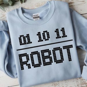 Könnte beinhalten: Hellblaues Sweatshirt mit dem Text "01 10 11 / ROBOT" in schwarzer Pixel-Schrift. Das Sweatshirt hat einen Rundhalsausschnitt und lange Ärmel. Das Gildan-Markenschild ist am Ausschnitt sichtbar.