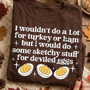 May include: A dark brown t-shirt featuring white text that reads I wouldn't do a Lot for turkey or ham but i would do some sketchy stuff for deviled eggs, accompanied by three illustrations of halved deviled eggs and small star icons.
