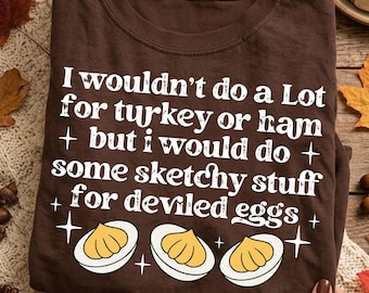 May include: A dark brown t-shirt featuring white text that reads I wouldn't do a Lot for turkey or ham but i would do some sketchy stuff for deviled eggs, accompanied by three illustrations of halved deviled eggs and small star icons.