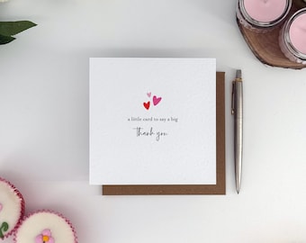 May include: A white card with the message "a little card to say a big Thank you" and three small hearts. The card sits on a brown backing, with a silver pen beside it. Cupcakes and candles are in the background.