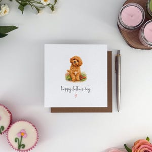 May include: A square greeting card with a brown border features a watercolour illustration of a golden-brown dog in a flower bed. The text "happy fathers day" is written below the dog, with a small heart symbol.