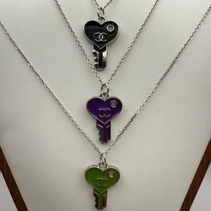 May include: Three silver necklaces with heart-shaped key pendants. The pendants are black, purple, and green with silver key details. Each pendant has a small circular accent and a double-C logo.