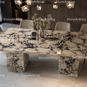 Op de afbeelding: Een rechthoekige eettafel met een gepolijst stenen oppervlak met een opvallend crème, beige en zwart marmerpatroon. De tafel wordt ondersteund door twee dikke, bijpassende stenen voetstukken en is omringd door vier beige gestoffeerde stoelen.