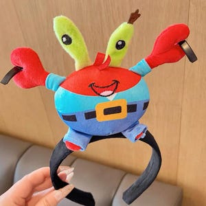 Puede incluir: Una diadema de felpa con un personaje de cangrejo de dibujos animados con pinzas rojas, cuerpo rojo, pantalones azules y ojos verdes. El cangrejo tiene una cara sonriente y está unido a una diadema negra.