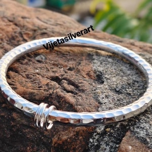 May include: A hammered silver bangle bracelet with a textured surface. The bracelet has a simple design with a small, decorative loop. The text "Vijetasilverart" is visible on the image.