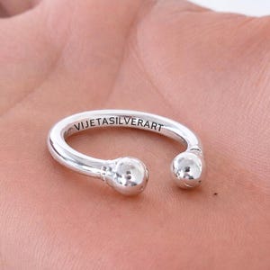 May include: A silver ring with a curved design and two ball-shaped ends. The ring has the text "VIJETASILVERART" engraved on the band. The ring is displayed on a light-coloured surface.