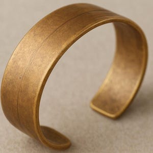 May include: A bronze cuff bracelet with a textured surface. The open-ended bracelet has a simple design, with a single groove along the top. The metal has a warm, antique finish. The bracelet is approximately 2.5 cm wide.