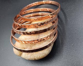 May include: A stack of seven copper bangles with a hammered texture. The bracelets are a warm, reddish-brown color and are resting on a light-colored stone against a dark background. The bangles are simple and elegant.