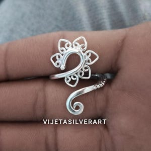 May include: A silver ring with a floral design. The ring has a flower-shaped top with detailed cutouts and a swirling band. The text "VIJETASILVERART" is visible. The ring's design is intricate and eye-catching.
