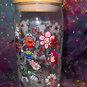 K&ouml;nnte beinhalten: Ein durchsichtiges Glas mit Bambusdeckel, das mit einem bunten Rundumdruck verschiedener Zeichentrickfiguren versehen ist, darunter ein rosa B&auml;r, ein gr&uuml;ner Weltraum-Ranger, eine kartoffelf&ouml;rmige Figur und ein Einhorn, umgeben von Blumen.