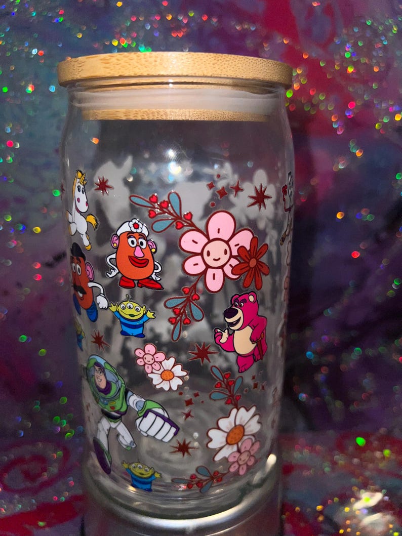 K&ouml;nnte beinhalten: Ein durchsichtiges Glas mit Bambusdeckel, das mit einem bunten Rundumdruck verschiedener Zeichentrickfiguren versehen ist, darunter ein rosa B&auml;r, ein gr&uuml;ner Weltraum-Ranger, eine kartoffelf&ouml;rmige Figur und ein Einhorn, umgeben von Blumen.