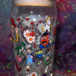 K&ouml;nnte beinhalten: Ein durchsichtiges Glas mit Bambusdeckel, das mit einem bunten Rundumdruck verschiedener Zeichentrickfiguren versehen ist, darunter ein Cowboy, ein Weltraum-Ranger, eine kartoffelf&ouml;rmige Figur, ein Dinosaurier, ein Schwein, eine Sch&auml;ferin und ein Einhorn.