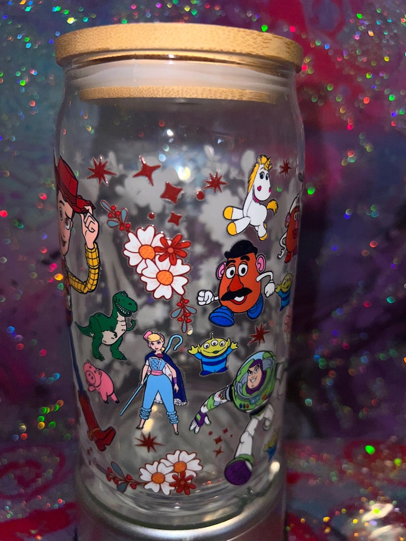 K&ouml;nnte beinhalten: Ein durchsichtiges Glas mit Bambusdeckel, das mit einem bunten Rundumdruck verschiedener Zeichentrickfiguren versehen ist, darunter ein Cowboy, ein Weltraum-Ranger, eine kartoffelf&ouml;rmige Figur, ein Dinosaurier, ein Schwein, eine Sch&auml;ferin und ein Einhorn.