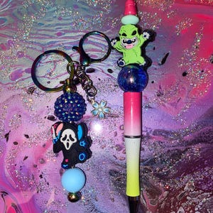 Op de afbeelding: Een aangepaste pen- en sleutelhanger set. De pen heeft een verloopontwerp met een groene karaktertop. De sleutelhanger heeft een zwarte karakterbedel, een blauwe kraal en een blauwe glitterbal. De set is ontworpen met een kleurrijk, glinsterend thema.