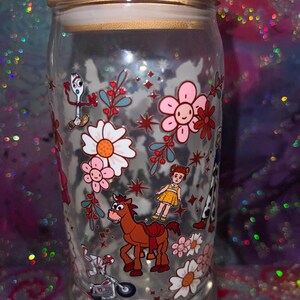 K&ouml;nnte beinhalten: Ein durchsichtiges Glas mit Bambusdeckel, das mit einem bunten Rundumdruck von Zeichentrickfiguren wie einer wei&szlig;en Gabel, einem braunen Pferd und einer kleinen Puppe sowie rosa und wei&szlig;en Blumen, roten Sternen und Zweigen verziert ist.