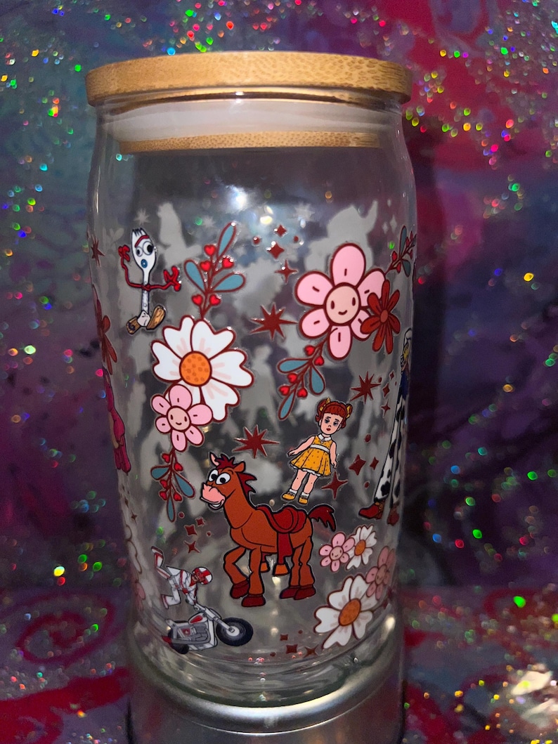 K&ouml;nnte beinhalten: Ein durchsichtiges Glas mit Bambusdeckel, das mit einem bunten Rundumdruck von Zeichentrickfiguren wie einer wei&szlig;en Gabel, einem braunen Pferd und einer kleinen Puppe sowie rosa und wei&szlig;en Blumen, roten Sternen und Zweigen verziert ist.