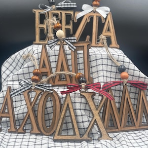 Può includere: Ornamenti in legno con lettere greche, decorati con perline e nastri decorativi. Le lettere sono di colore marrone scuro, con nastri a scacchi bianchi e neri, rossi e neri e bianchi. Gli ornamenti sono esposti su un tessuto a scacchi.