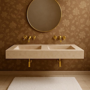 May include: A modern bathroom sink with two basins, made of light beige stone. The sink has gold-colored faucets and drain pipes. A round gold mirror hangs above the sink, and the wall is covered in a floral wallpaper. A white rug is on the floor.