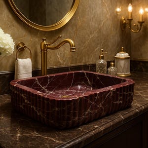 Puede incluir: Un lavabo de ba&ntilde;o de m&aacute;rmol burdeos con vetas blancas, rectangular con dise&ntilde;o acanalado. Grifo, espejo y apliques dorados. Un jarr&oacute;n dorado contiene flores blancas. Un conjunto elegante para un ba&ntilde;o con un toque de lujo.