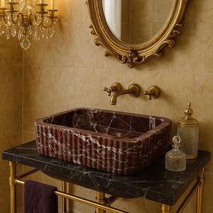 Puede incluir: Un lavabo rectangular de m&aacute;rmol burdeos acanalado se asienta sobre un tocador de m&aacute;rmol negro con patas doradas. Un espejo ovalado con marco dorado y una l&aacute;mpara de ara&ntilde;a est&aacute;n montados en la pared de azulejos beige. Botellas de vidrio decorativas y una toalla completan la escena del ba&ntilde;o.