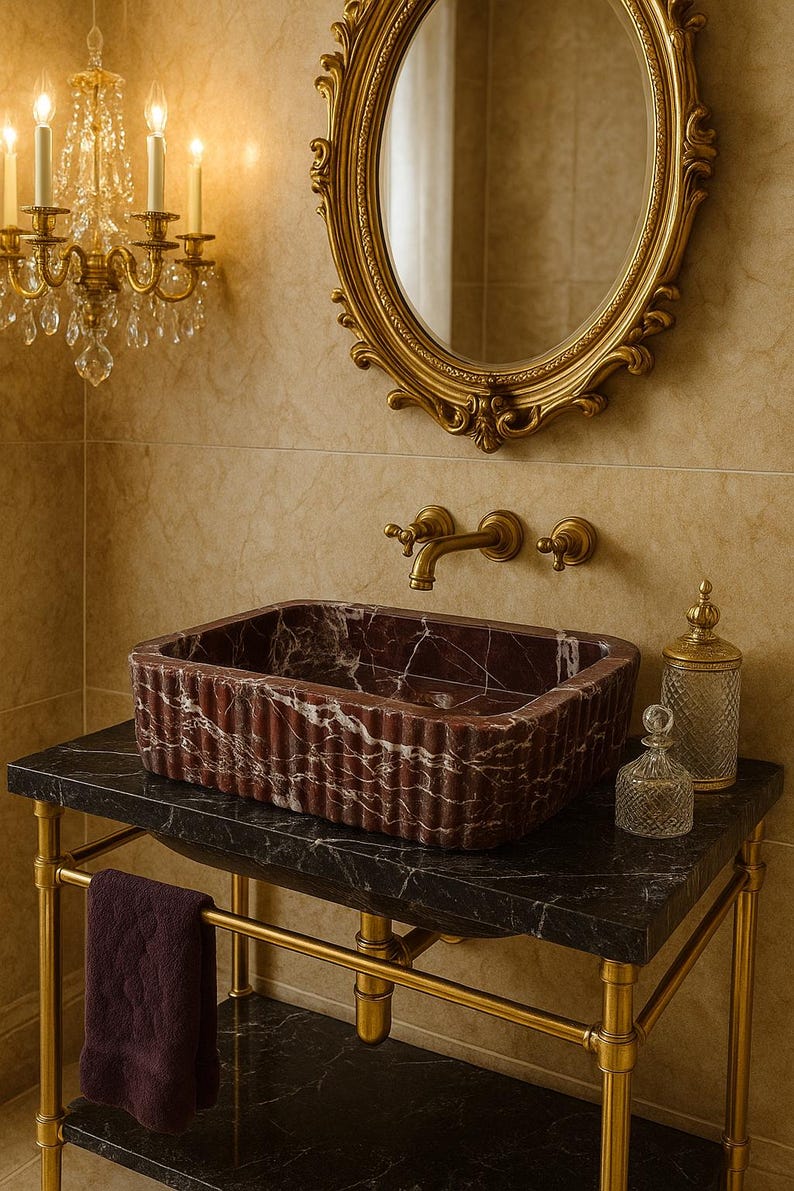 Puede incluir: Un lavabo rectangular de m&aacute;rmol burdeos acanalado se asienta sobre un tocador de m&aacute;rmol negro con patas doradas. Un espejo ovalado con marco dorado y una l&aacute;mpara de ara&ntilde;a est&aacute;n montados en la pared de azulejos beige. Botellas de vidrio decorativas y una toalla completan la escena del ba&ntilde;o.