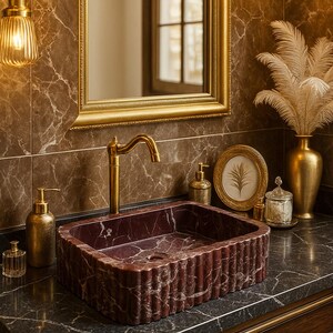 Puede incluir: Un lujoso mueble de ba&ntilde;o con un lavabo rectangular de m&aacute;rmol burdeos con un dise&ntilde;o estriado. El lavabo est&aacute; sobre una encimera oscura con accesorios dorados, un espejo con marco dorado y detalles dorados decorativos.