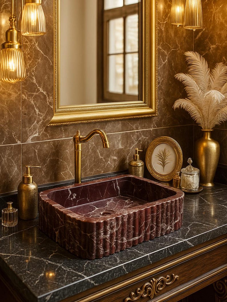 Puede incluir: Un lujoso mueble de ba&ntilde;o con un lavabo rectangular de m&aacute;rmol burdeos con un dise&ntilde;o estriado. El lavabo est&aacute; sobre una encimera oscura con accesorios dorados, un espejo con marco dorado y detalles dorados decorativos.