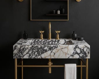 Puede incluir: Un lujoso mueble de baño con un lavabo de mármol con vetas negras, sobre una estructura metálica dorada. El mueble incluye un grifo dorado y apliques a juego, un espejo y una toalla blanca. Una alfombra de baño de color claro está en el suelo.