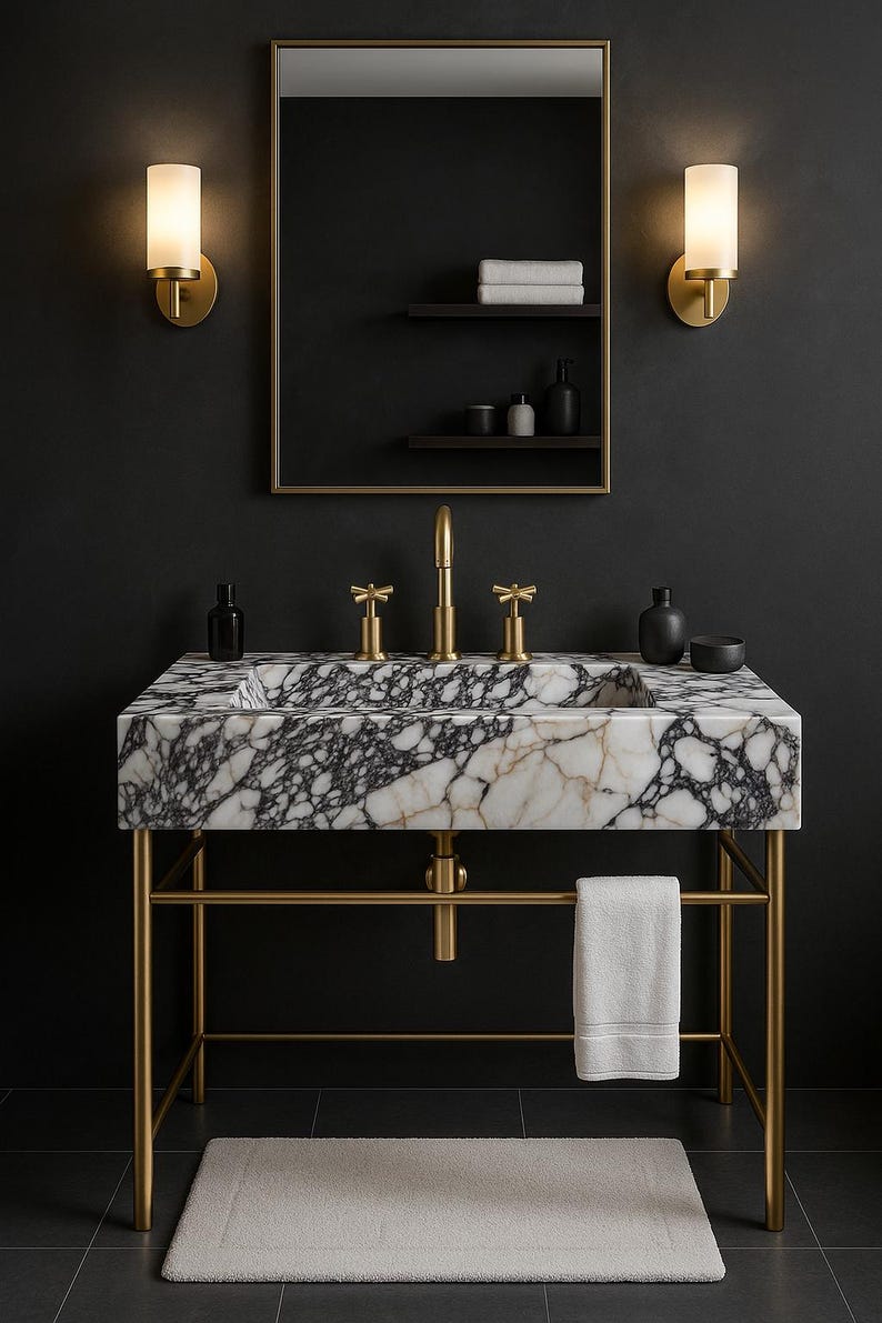 Op de afbeelding: Een luxe badkamermeubel met een marmeren wastafel met zwarte adering, geplaatst op een goudkleurig metalen frame. De wastafel heeft een gouden kraan en bijpassende wandkandelaars, een spiegel en een witte handdoek. Een lichtgekleurde badmat ligt op de vloer.