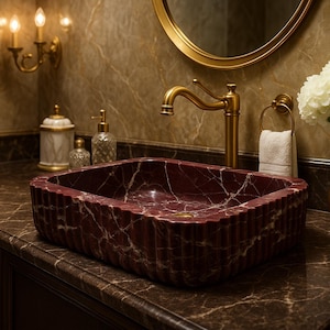 Puede incluir: Un lavabo rectangular de m&aacute;rmol burdeos con vetas blancas se encuentra sobre una encimera oscura. Se ven un grifo, un espejo y apliques dorados con velas encendidas. Un jarr&oacute;n dorado contiene flores blancas.