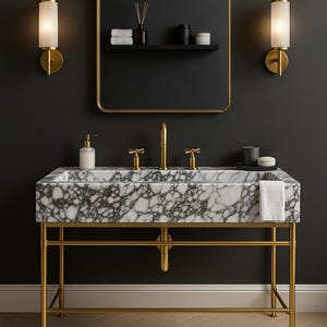 Op de afbeelding: Een luxe badkamermeubel met een marmeren wastafel en gouden armaturen. De wastafel wordt ondersteund door een gouden metalen frame en een goudomrande spiegel hangt erboven. Twee wandkandelaars flankeren de spiegel en een grijs tapijt ligt op de vloer.