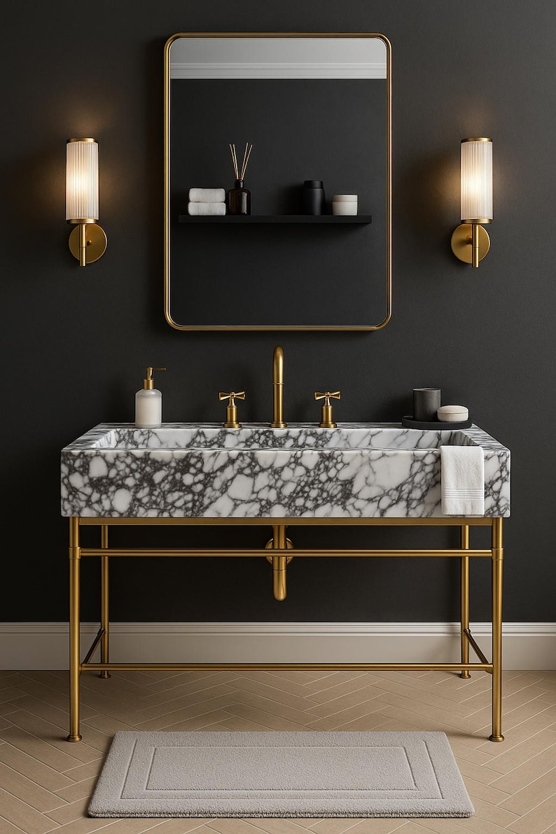 Op de afbeelding: Een luxe badkamermeubel met een marmeren wastafel en gouden armaturen. De wastafel wordt ondersteund door een gouden metalen frame en een goudomrande spiegel hangt erboven. Twee wandkandelaars flankeren de spiegel en een grijs tapijt ligt op de vloer.
