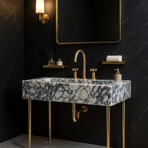 Op de afbeelding: Een moderne badkamermeubel met een marmeren wastafel met gouden kranen en poten. De wastafel staat tegen een zwarte marmeren muur, met een goudomrande spiegel en een wandlamp. Een beige tapijt ligt op de vloer. De wastafel is ongeveer 100 cm lang.