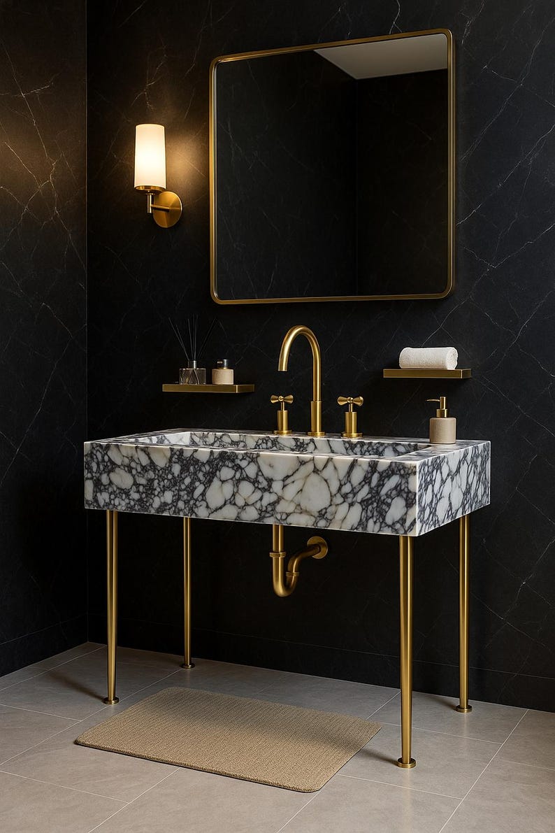 Op de afbeelding: Een moderne badkamermeubel met een marmeren wastafel met gouden kranen en poten. De wastafel staat tegen een zwarte marmeren muur, met een goudomrande spiegel en een wandlamp. Een beige tapijt ligt op de vloer. De wastafel is ongeveer 100 cm lang.