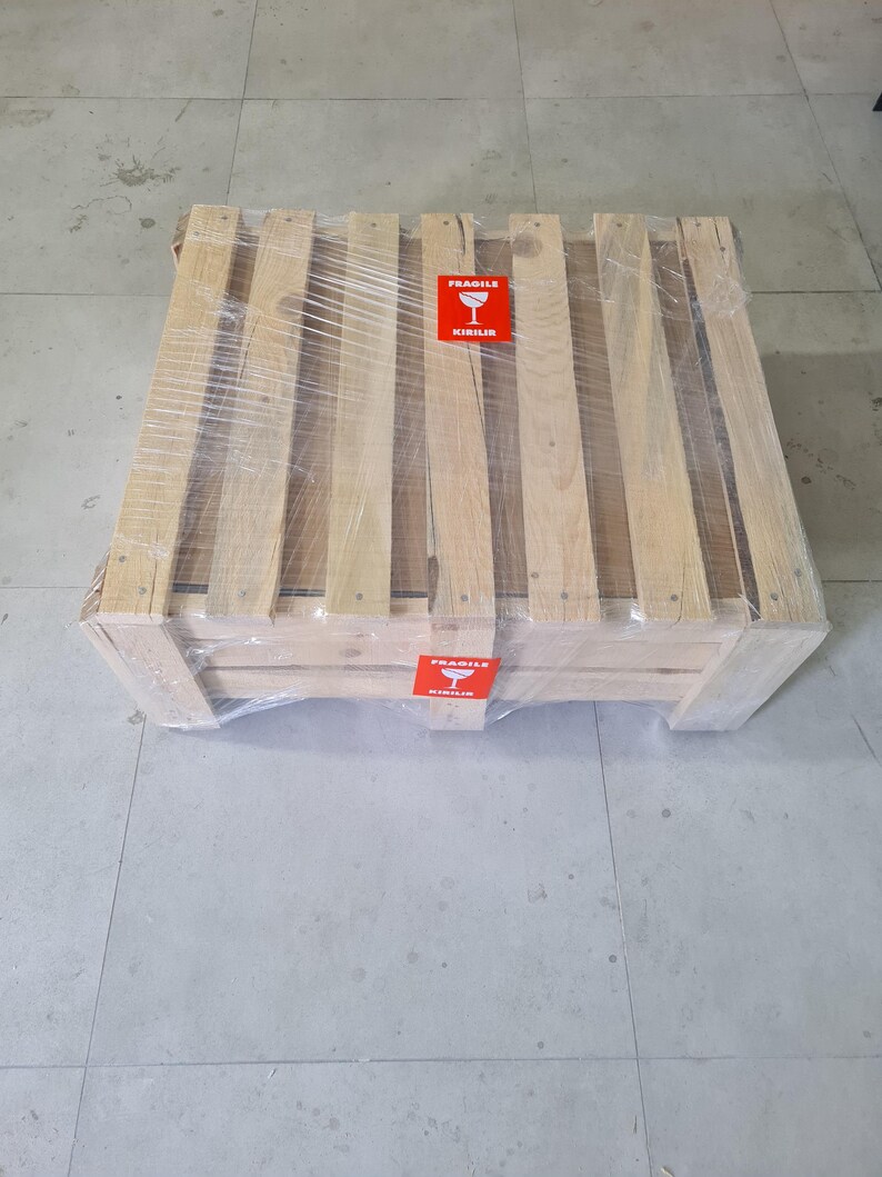 Puede incluir: Un palet de env&iacute;o de madera envuelto en pl&aacute;stico transparente. El palet tiene varias lamas y soportes de madera. Se ven dos pegatinas rojas de "Fr&aacute;gil".
