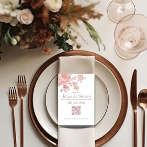 May include: A rectangular white wedding place card featuring a soft pink floral illustration at the top, the names Julija & Stevan, the date OKT 27 2028, and a square QR code at the bottom, resting on a folded beige napkin atop a white and copper plate.