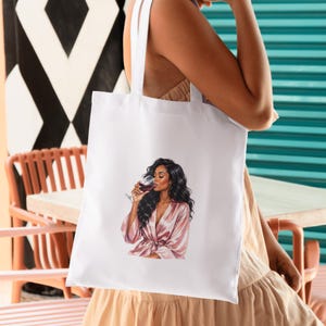 May include: White tote bag featuring an illustration of a person in a pink robe drinking wine. The person has long, dark wavy hair. The tote bag has white straps and is held by a person wearing a beige dress.