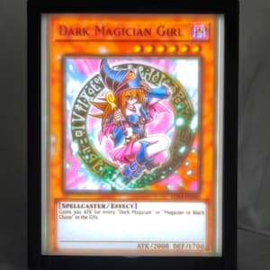 May include: A framed card featuring the "Dark Magician Girl" from the Yu-Gi-Oh! trading card game. The card has a colorful illustration of a female character with text at the bottom, including ATK/2000 DEF/1700.
